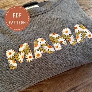 May include: A gray sweatshirt with the word "MAMA" embroidered in a daisy floral pattern. The sweatshirt has a textured appearance and a round, ribbed neckline. A circular sticker with the text "PDF PATTERN" is visible.