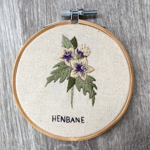 May include: Embroidered hoop art featuring a cluster of yellow and purple flowers with green leaves. The text "HENBANE" is embroidered below the flowers.