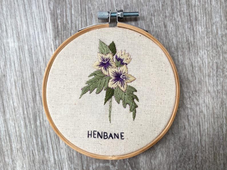 May include: Embroidered hoop art featuring a cluster of yellow and purple flowers with green leaves. The text "HENBANE" is embroidered below the flowers.