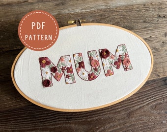 May include: A white embroidery hoop with the word "MUM" embroidered in red thread. The letters are filled with a floral pattern in shades of pink, red, and green.