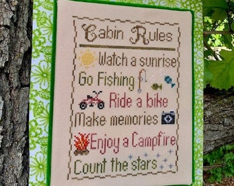 May include: A green and white framed sign with the text "Cabin Rules" and a list of rules for enjoying a cabin holiday: watch a sunrise, go fishing, ride a bike, make memories, enjoy a campfire, and count the stars.