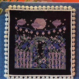 Peut inclure: Broderie au point de croix représentant une scène de jardin nocturne. Le motif comprend une clôture, des fleurs et une guirlande lumineuse sous une lune et des étoiles. L'œuvre est encadrée d'une bordure en tissu bleu et d'une garniture en pompons blancs.