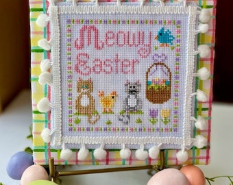 Puede incluir: Un bordado de punto de cruz blanco con un borde a cuadros rosa y blanco y pompones. El bordado presenta el texto "Meowy Easter" con un gato, un pollito y una cesta de huevos.