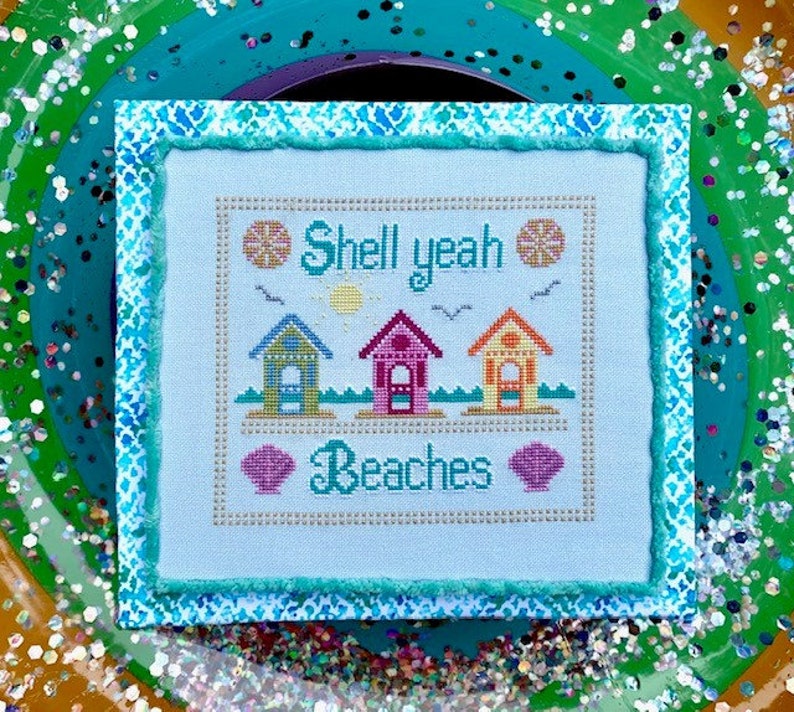 May include: A cross-stitch design featuring three beach houses, a sun, two seashells, and the text "Shell yeah Beaches". The design is framed with a blue and white patterned border.