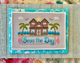 May include: A cross-stitch design framed on a wooden board with a blue and white background. The design features a beach house with palm trees and the text "Seas the Day".