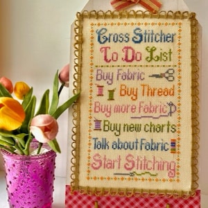 Puede incluir: Un sampler de punto de cruz con fondo beige y marco dorado. El sampler dice "Cross Stitcher To Do List" e incluye una lista de tareas relacionadas con el punto de cruz, como "Comprar tela", "Comprar hilo", "Comprar más tela", "Comprar nuevos gráficos", "Hablar de tela" y "Empezar a bordar".