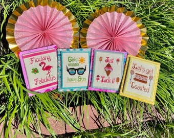 May include: Four framed cross-stitch designs with summer themes. The designs are "Flocking Fabulous" with a pink flamingo, "Girls wanna have Sun" with sunglasses and a sun, "Just Lick it" with an ice cream cone, and "Let's get Toasted" with a slice of toast and a lollipop.