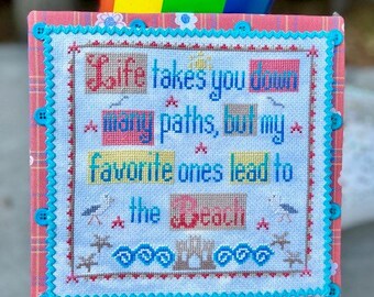 Puede incluir: Un sampler de punto de cruz con un borde azul y blanco y un fondo de cuadros rosa y blanco. El sampler presenta el texto "Life takes you down many paths, but my favorite ones lead to the beach" con una escena de playa debajo.