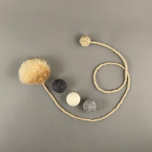 Könnte beinhalten: Ein brauner und weißer Fellpompon, der an einem Naturjute-Seil mit einem Knoten am Ende befestigt ist. Es gibt vier Filzkugeln in verschiedenen Farben: schwarz, weiß und zwei grau.