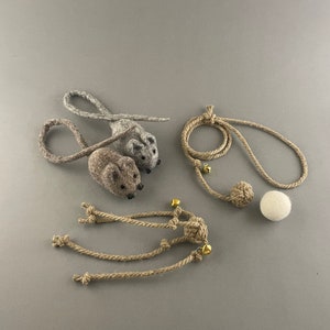 Może przedstawiać: Dwie szare myszy z filcu z brązową myszą z filcu i brązową sznurkową zabawką z dzwonkiem i białą piłką na szarym tle.