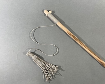 May include: A wooden cat toy with a light brown, striped stick, wrapped with gray twine. A gray tassel is attached to the end of a gray string. The toy is set against a gray background.