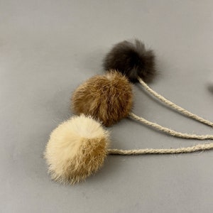 May include: Three fluffy cat toys in cream, brown, and dark gray. Each toy is a small, round ball of fur attached to a natural-colored rope. The toys are arranged diagonally against a gray background, showcasing their textures and colors.