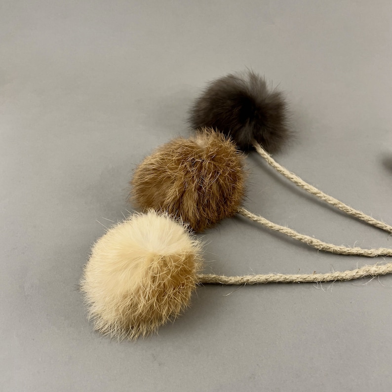 May include: Three fluffy cat toys in cream, brown, and dark gray. Each toy is a small, round ball of fur attached to a natural-colored rope. The toys are arranged diagonally against a gray background, showcasing their textures and colors.