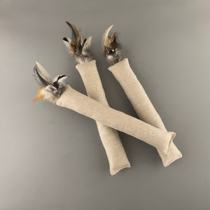 May include: Three beige cat toys with feathers. Each toy is a long, cylindrical tube made of a natural fabric with a feather sticking out of one end.