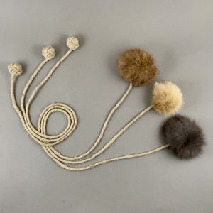 May include: A collection of cat toys featuring natural rope and fluffy fur balls in shades of brown, tan, and gray. The toys have long rope handles and knotted ends, designed for interactive play. The background is a solid gray.
