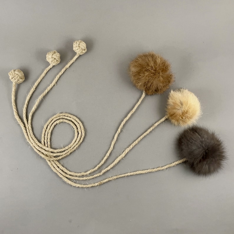 May include: A collection of cat toys featuring natural rope and fluffy fur balls in shades of brown, tan, and gray. The toys have long rope handles and knotted ends, designed for interactive play. The background is a solid gray.
