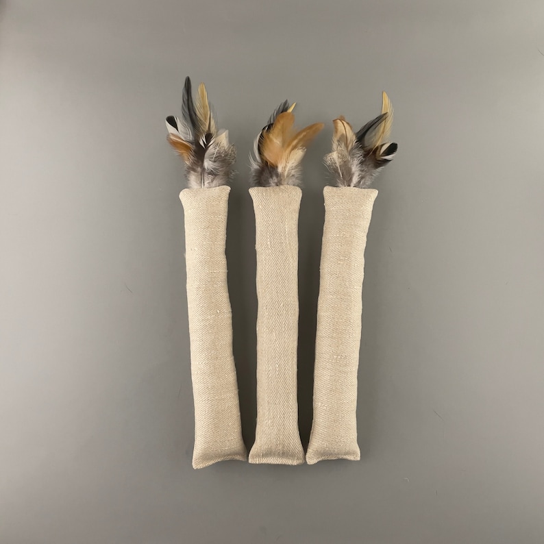 May include: Three beige cat toys with feathers. Each toy is a long, narrow tube made of natural linen fabric with a cluster of brown, black, and white feathers at the top.