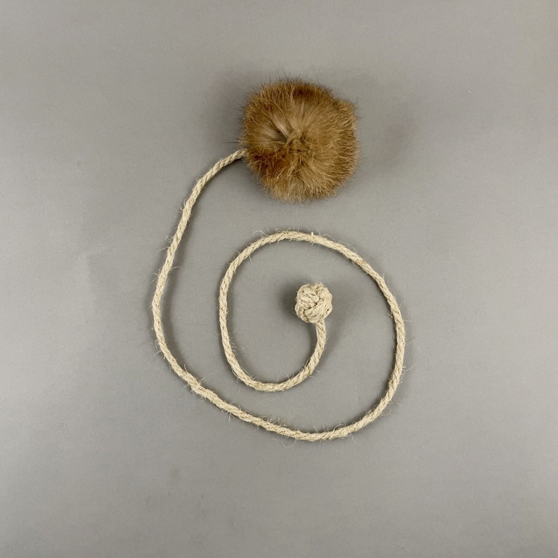 May include: A cat toy featuring a brown, fluffy ball attached to a natural-colored rope. The rope is coiled into a spiral shape, with a knotted end. The toy is designed for feline play and enrichment.