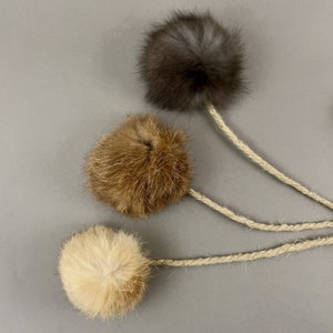 May include: Three cat toys with fluffy, round, fur balls in shades of brown, tan, and gray. Each ball is attached to a natural-colored rope. The toys are arranged against a gray background.