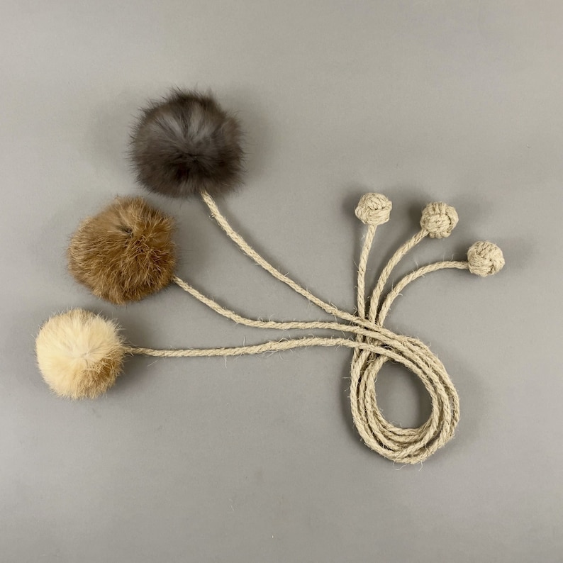 May include: A collection of cat toys featuring fluffy, round balls in shades of gray, brown, and cream, attached to natural rope. The toys are designed with a playful aesthetic, perfect for feline entertainment.
