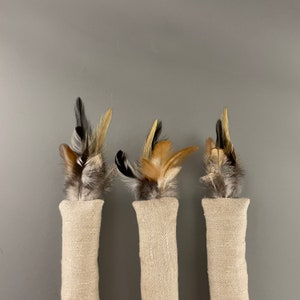 May include: Three beige fabric cat toys with different colored feathers. The feathers are brown, black, and white.
