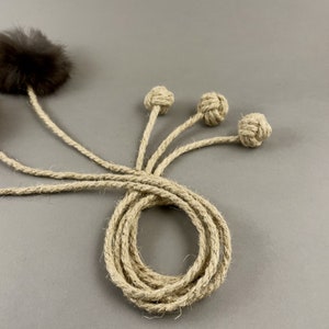 May include: A cat toy featuring a brown fur ball and three knotted rope balls attached to a coiled rope. The rope and knots are a natural beige color, set against a gray background. This toy is designed for feline entertainment.