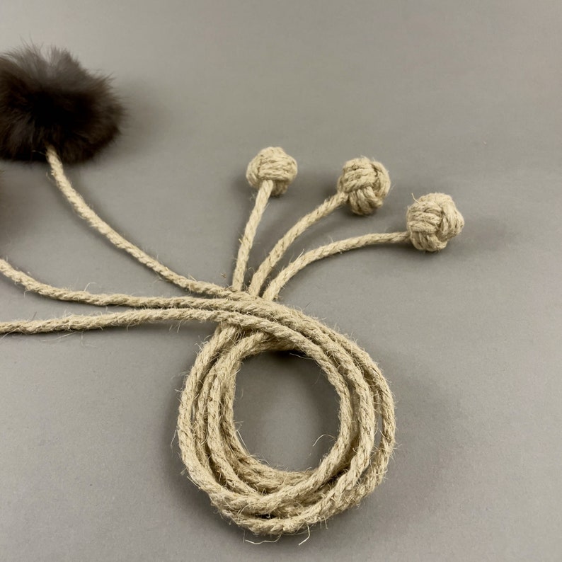 May include: A cat toy featuring a brown fur ball and three knotted rope balls attached to a coiled rope. The rope and knots are a natural beige color, set against a gray background. This toy is designed for feline entertainment.