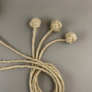 May include: Three decorative rope knot accents with long rope stems, arranged against a gray background. The knots are spherical and the rope is a natural beige color. The rope stems extend from the knots and are intertwined.
