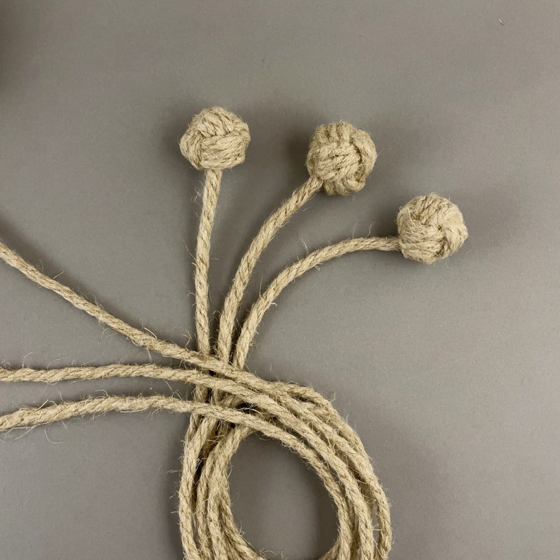 May include: Three decorative rope knot accents with long rope stems, arranged against a gray background. The knots are spherical and the rope is a natural beige color. The rope stems extend from the knots and are intertwined.
