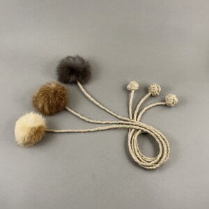May include: Cat toys featuring three fluffy pom-poms in cream, brown, and gray, attached to twine strings. The image also shows four knotted twine toys. The toys are arranged on a gray background.