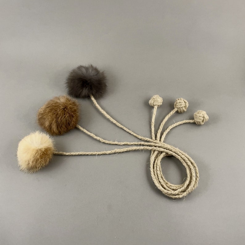 May include: Cat toys featuring three fluffy pom-poms in cream, brown, and gray, attached to twine strings. The image also shows four knotted twine toys. The toys are arranged on a gray background.