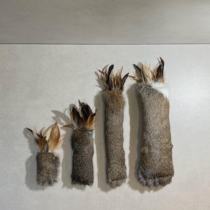May include: Four handmade pet toys made from natural materials. Each toy is a tube of fur with a cluster of brown and tan feathers at the top. The toys vary in size, with the largest being approximately 25 cm long.