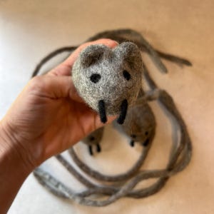 Może przedstawiać: Zestaw trzech małych, okrągłych, szarych myszek z wełny filcowanej igłą, z czarnymi wyszywanymi oczami i czarnymi filcowymi łapkami. Każda mysz ma długi, cienki, szary filcowy ogon, który owija się wokół grupy, tworząc zestaw figurek zwierząt.
