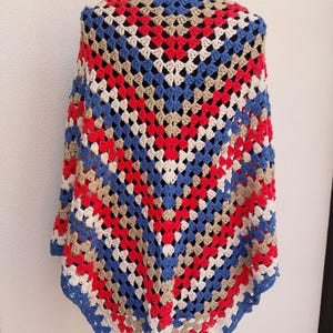Puede incluir: Un chal de ganchillo con un diseño de granny square. El chal presenta una vibrante mezcla de cuadrados rojos, blancos, azules y beige, creando un patrón geométrico. Los bordes están acabados con un borde azul.