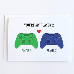 May include: A white card with the text "You're my Player 2" and a red heart.  Two game controllers are shown, one green and one blue, with the text "Player 1" and "Player 2" below them.