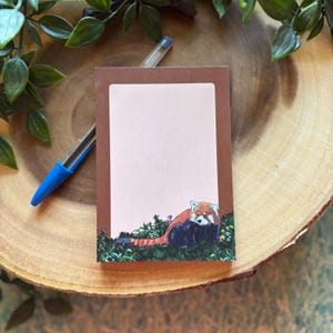 May include: A rectangular notepad with a brown border and a light pink interior. The bottom of the notepad features a red panda illustration in a grassy area with flowers. A blue pen rests on a wooden surface.