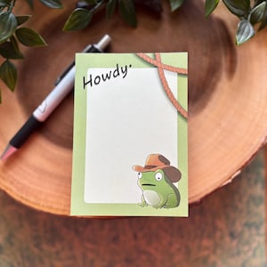 May include: A notepad with a light green border and the word "Howdy" in black script. A cartoon frog wearing a cowboy hat is in the lower right corner. A rope design is at the top. A pen is in the background.
