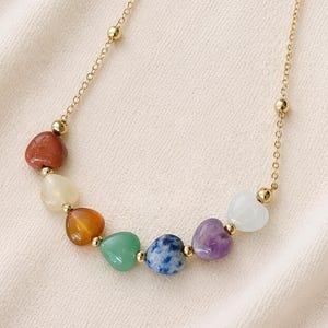 May include: A gold-colored chain necklace featuring heart-shaped gemstones in a rainbow of colors. The gemstones include red, yellow, orange, green, blue, purple, and white. The necklace has small gold beads interspersed along the chain.