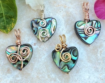 May include: Four heart-shaped pendants with iridescent abalone shells. Each pendant has a unique wire-wrapped design in silver, copper, or gold-colored metal. The pendants are displayed on a light-colored surface with floral accents.