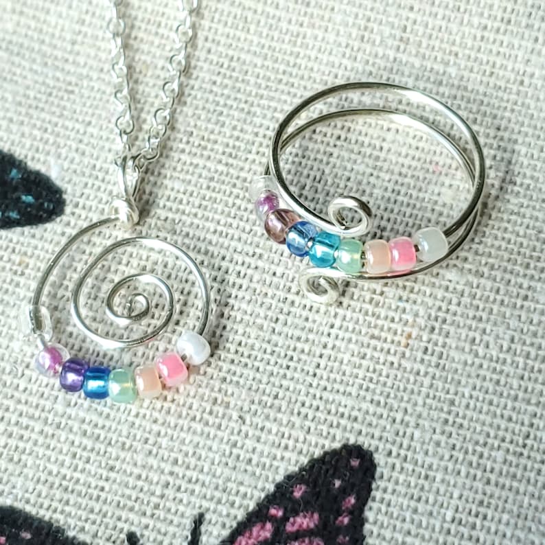 May include: A silver necklace and ring set with a spiral pendant and a beaded band. The beads are multi-colored and include pink, blue, green, and purple.