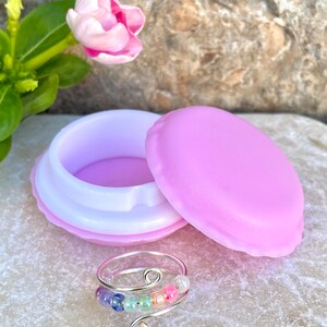 May include: A rainbow beaded ring with a silver wire design, displayed in a purple macaron-shaped box.