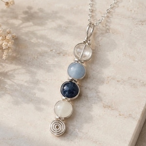 May include: A silver necklace featuring a pendant with four gemstone beads in clear, light blue, dark blue, and white. The pendant has a spiral design at the bottom and is strung on a delicate silver chain. The necklace is displayed on a light-colored surface.