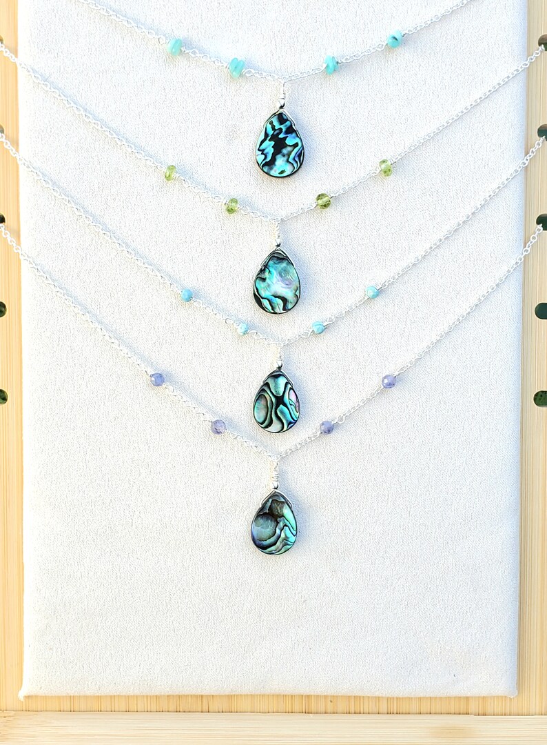 May include: Five silver necklaces with abalone shell pendants and various colored beads. The necklaces are displayed on a wooden background.