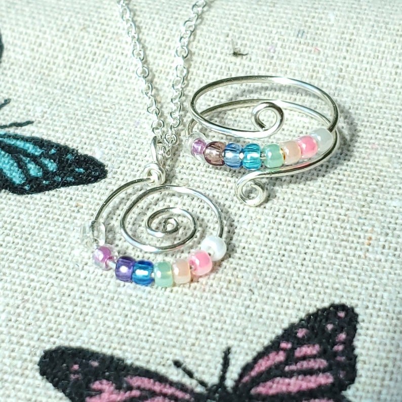 May include: A silver necklace and ring set with a spiral design and colorful beads. The beads are in shades of pink, blue, green, and purple.