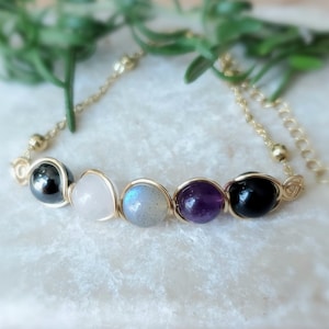 May include: A gold chain bracelet with five gemstone beads. The beads are black, white, grey, purple, and black. The bracelet is on a white surface.