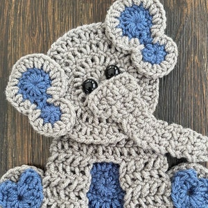 May include: Crocheted gray elephant with blue accents. The elephant has a heart-shaped nose and large ears. The text "Dark Country Blue" is written below the elephant.