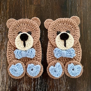 May include: Two handmade crochet teddy bears. Each bear has a brown body, cream-colored face, black nose, and blue bow tie and feet. The bears are displayed on a dark wood surface.