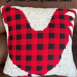 May include: A red and black buffalo plaid crocheted pillow in the shape of a rooster. The pillow is on a brown leather couch. The text "Get Hooked Crochet by Amanda Schmitt" is at the top of the image.