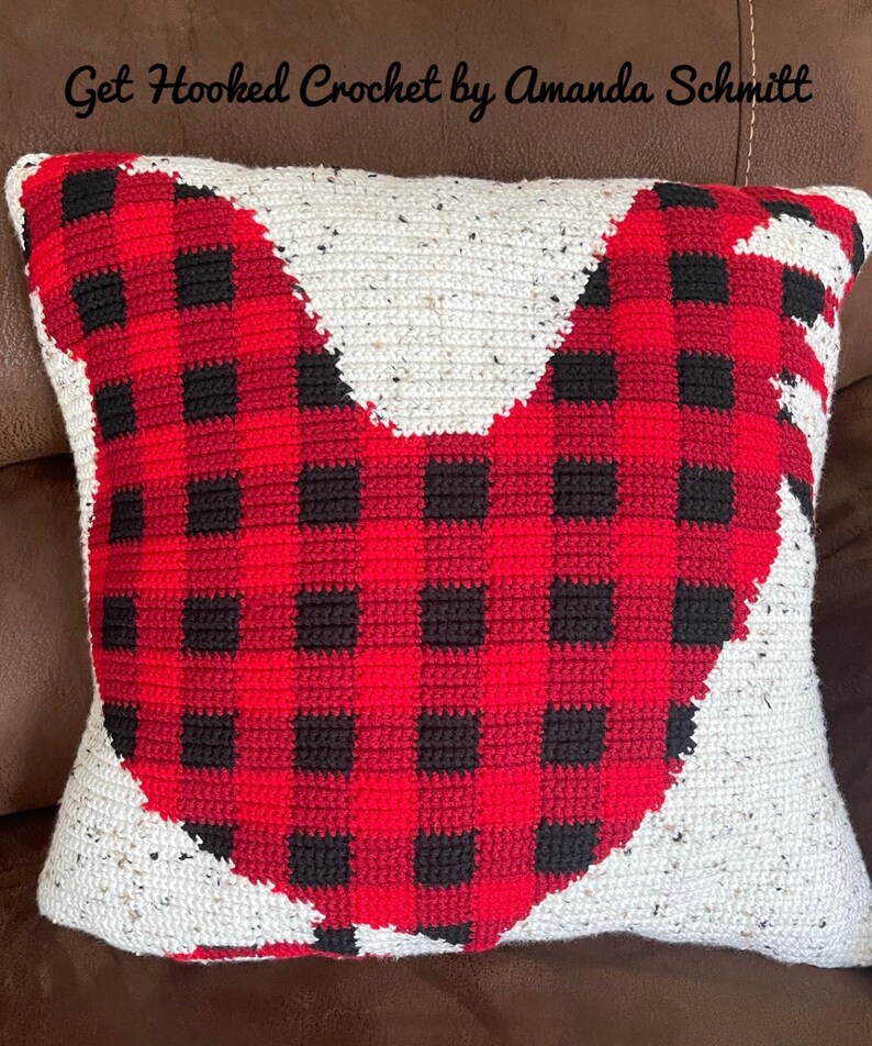 May include: A red and black buffalo plaid crocheted pillow in the shape of a rooster. The pillow is on a brown leather couch. The text "Get Hooked Crochet by Amanda Schmitt" is at the top of the image.