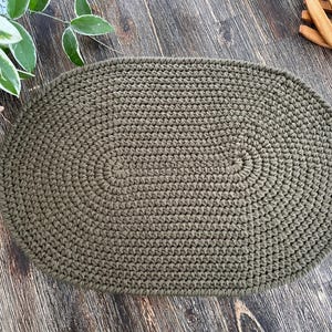 May include: A green oval crochet rug with a textured surface. The rug is made of thick yarn and has a simple, elegant design.